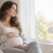 Pregnant woman sitting quietly by a window with one hand resting on her belly