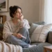 Woman sitting on a sofa by a window, holding a pregnancy test in a quiet living room.
