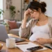 Pregnant woman sitting at a table at home with a laptop, looking stressed and tired while resting one hand on her forehead and the other on her belly.